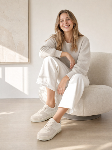 Woman wearing cream sneakers with white trousers, seated on a rounded chair.